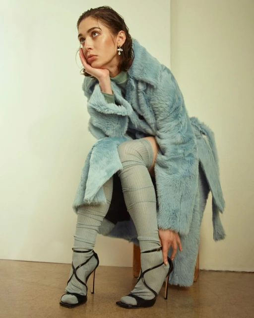 Woman in a light blue faux fur coat sits on a stool in a studio, wearing thigh-high socks and black high-heeled strappy shoes, looking off-camera.