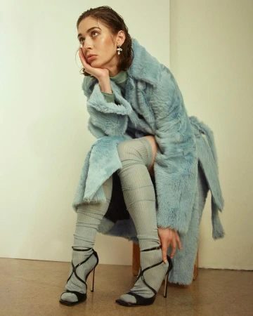 Woman in a light blue faux fur coat sits on a stool in a studio, wearing thigh-high socks and black high-heeled strappy shoes, looking off-camera.