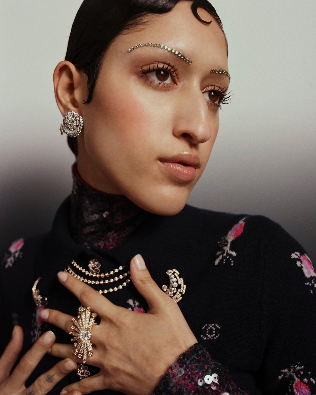 Close-up of an adult female model with crystal embellishments in hair and on eyebrows wearing a black floral-patterned jacket and jewelry.