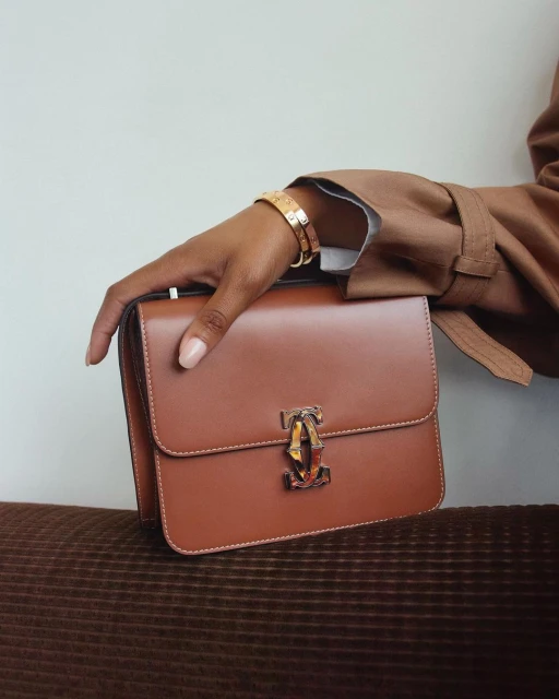 Brown leather purse with gold hardware held by a woman with tan skin and gold bracelets, set against a neutral backdrop.