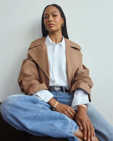 Black woman with braided hair wearing a tan jacket, white button-up shirt, and blue jeans by Emman Montalvan