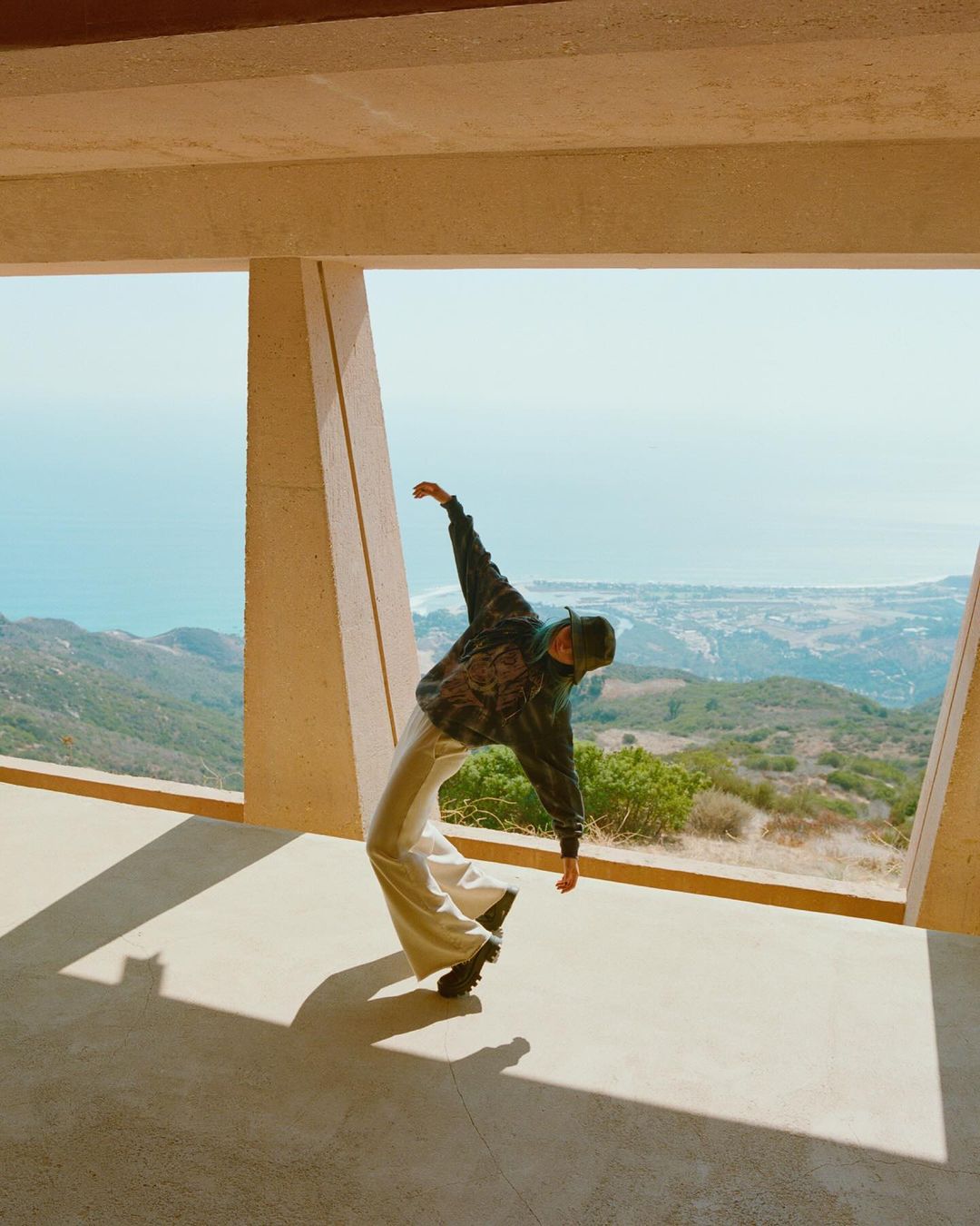 An adult dances near a window overlooking a distant mountain range and ocean under a clear sky in natural daylight.