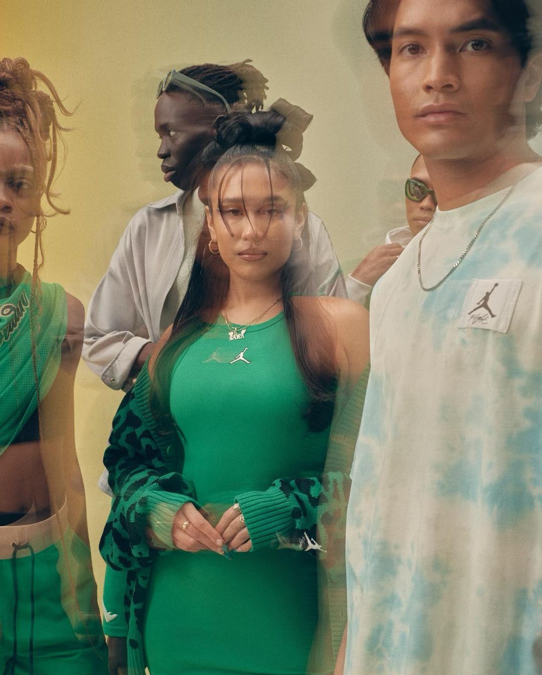 A group of five diverse young adults pose indoors, wearing contemporary apparel with brand logos, against a neutral yellow background.