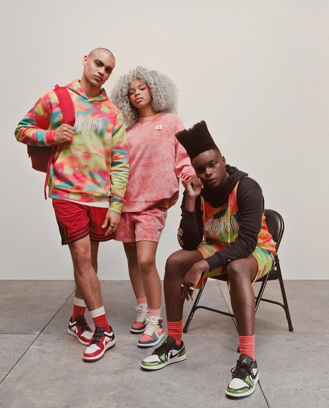 Three adult models wear tie-dye sportswear and sneakers in a studio setting. The models are styled with contemporary hair and poses.