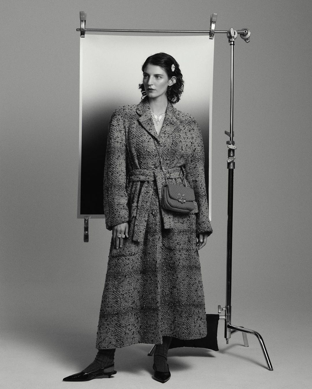 A woman in a long coat stands in a photo studio with a backdrop, accessorized with a small purse, jewelry, and black shoes. The image is black and white.