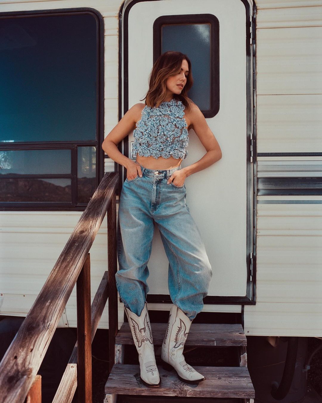 A woman in a floral crop top, jeans, and cowboy boots poses on the steps of a camper in an outdoor setting with mountains visible.
