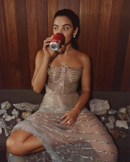 Adult woman seated, drinking from a red can, wearing a strapless sheer gown with decorative stones at hem and wood panel background.