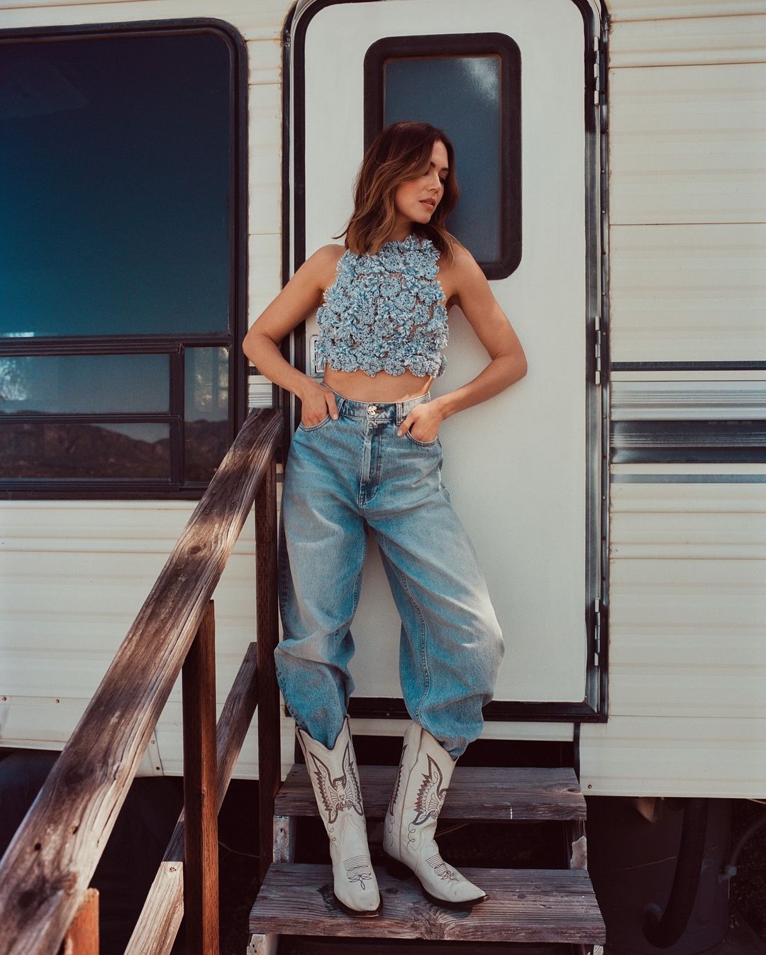 A brunette woman poses in front of a camper wearing a floral top, jeans, and cowboy boots on a sunny day with her hands in her pockets.