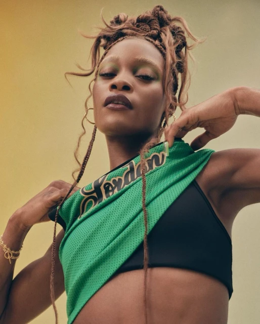 Adult female poses in green Jordan basketball jersey with black sports bra against yellow backdrop in a low-angle medium shot.