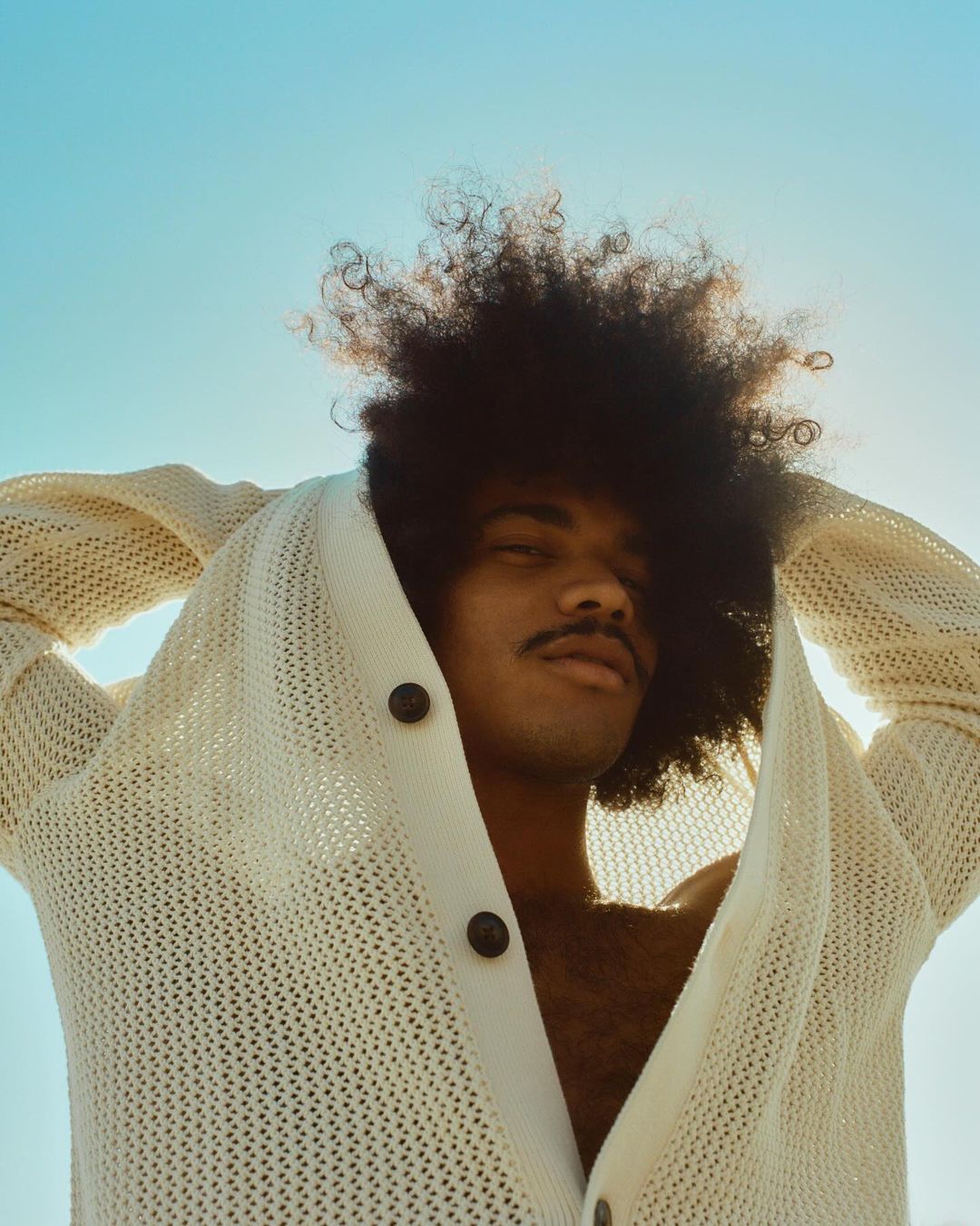 Close-up portrait of a young adult man with an afro, wearing a knit cardigan, against a clear blue sky in natural light.