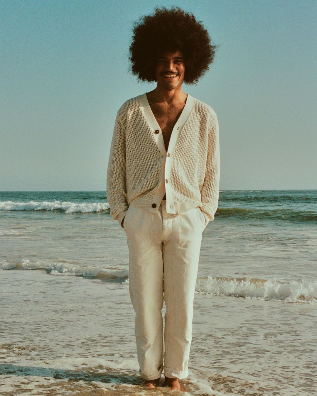 A smiling man with a large afro stands in the ocean wearing a cream cardigan and pants. The scene is naturally lit.