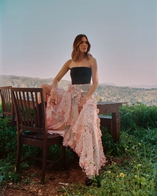 Mandy Moore sits on a chair outdoors, wearing a strapless top and a long, floral embellished skirt in a scenic, soft lighting.