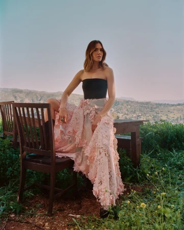 Mandy Moore sits on a chair outdoors, wearing a strapless top and a long, floral embellished skirt in a scenic, soft lighting.