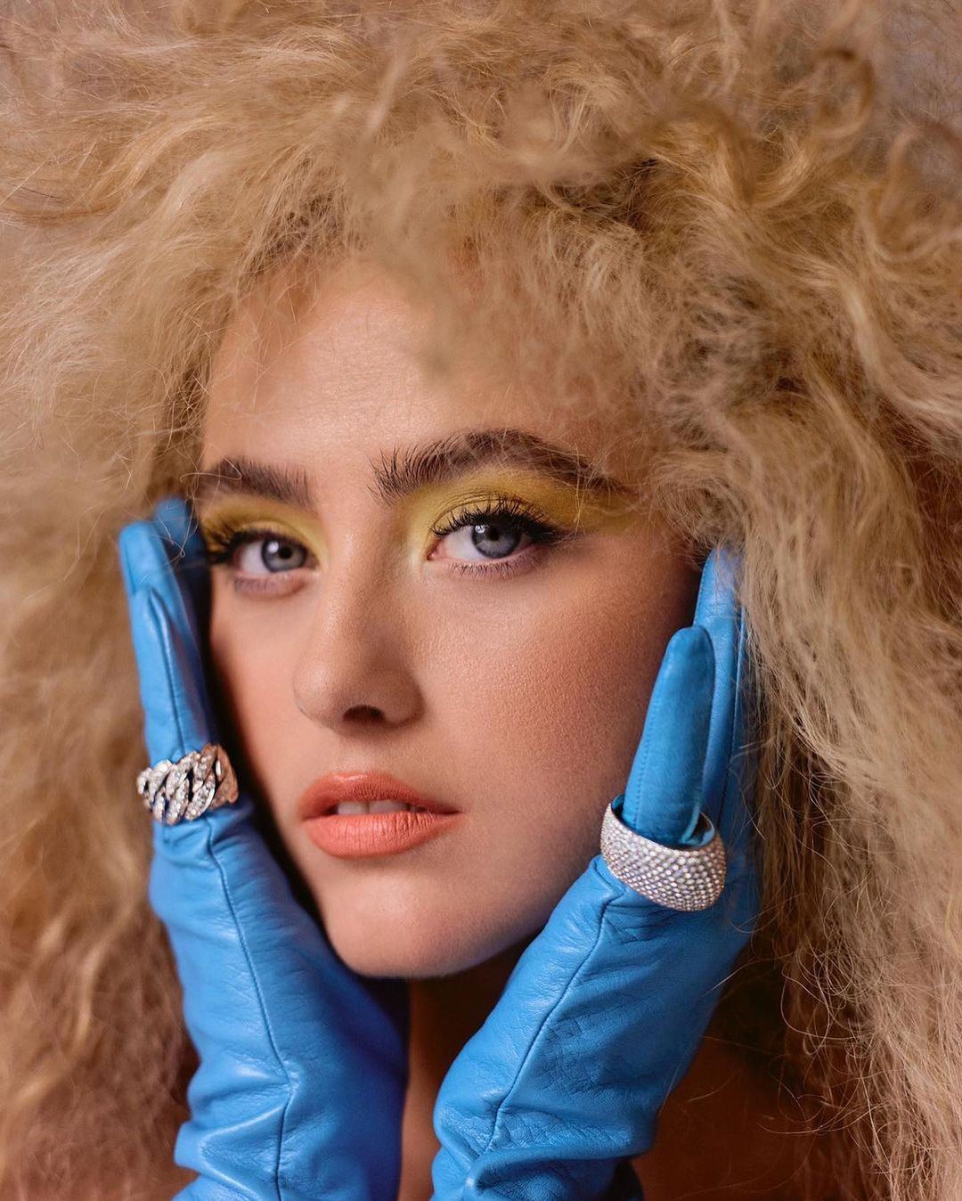 Close-up portrait of a woman with voluminous blonde hair wearing blue leather gloves framing her face, photographed by Emman Montalvan.