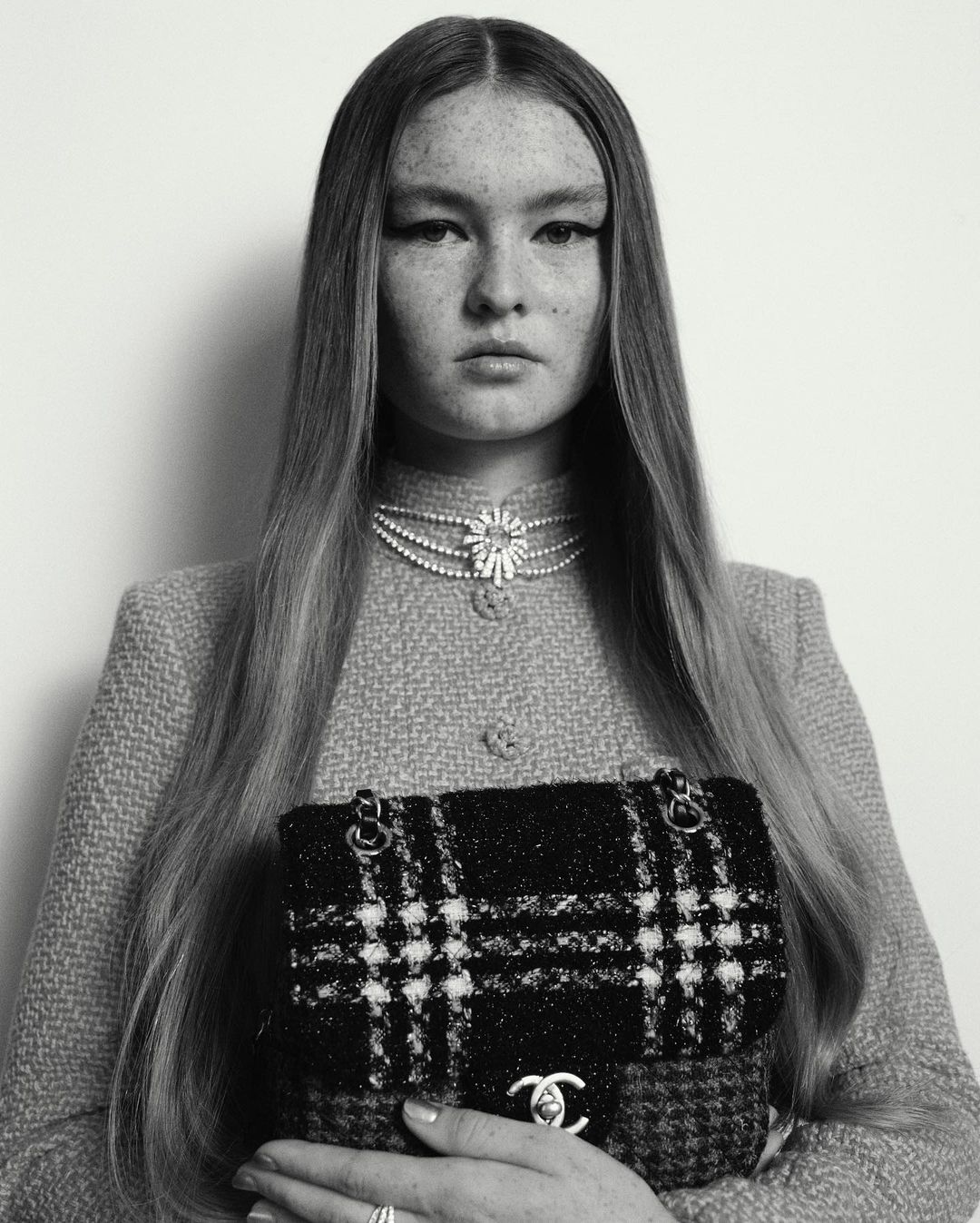 A fashion portrait of a young woman with freckles wearing a tweed jacket and pearl necklace, holding a plaid Chanel handbag in black and white.