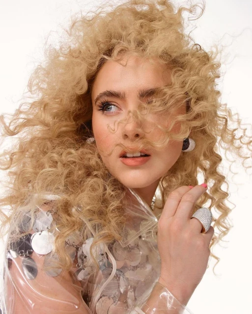 Blonde woman with curly hair wearing a clear sequined jacket in a studio portrait against a white background, beauty and fashion shoot.