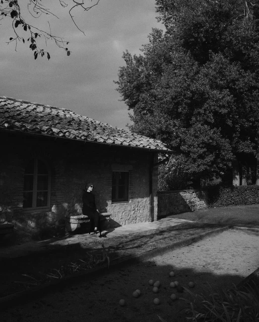 Black and white shot of an adult female in black clothing sitting on a stone bench outside a stone building in a yard.