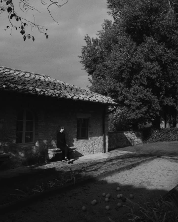 Black and white shot of an adult female in black clothing sitting on a stone bench outside a stone building in a yard.