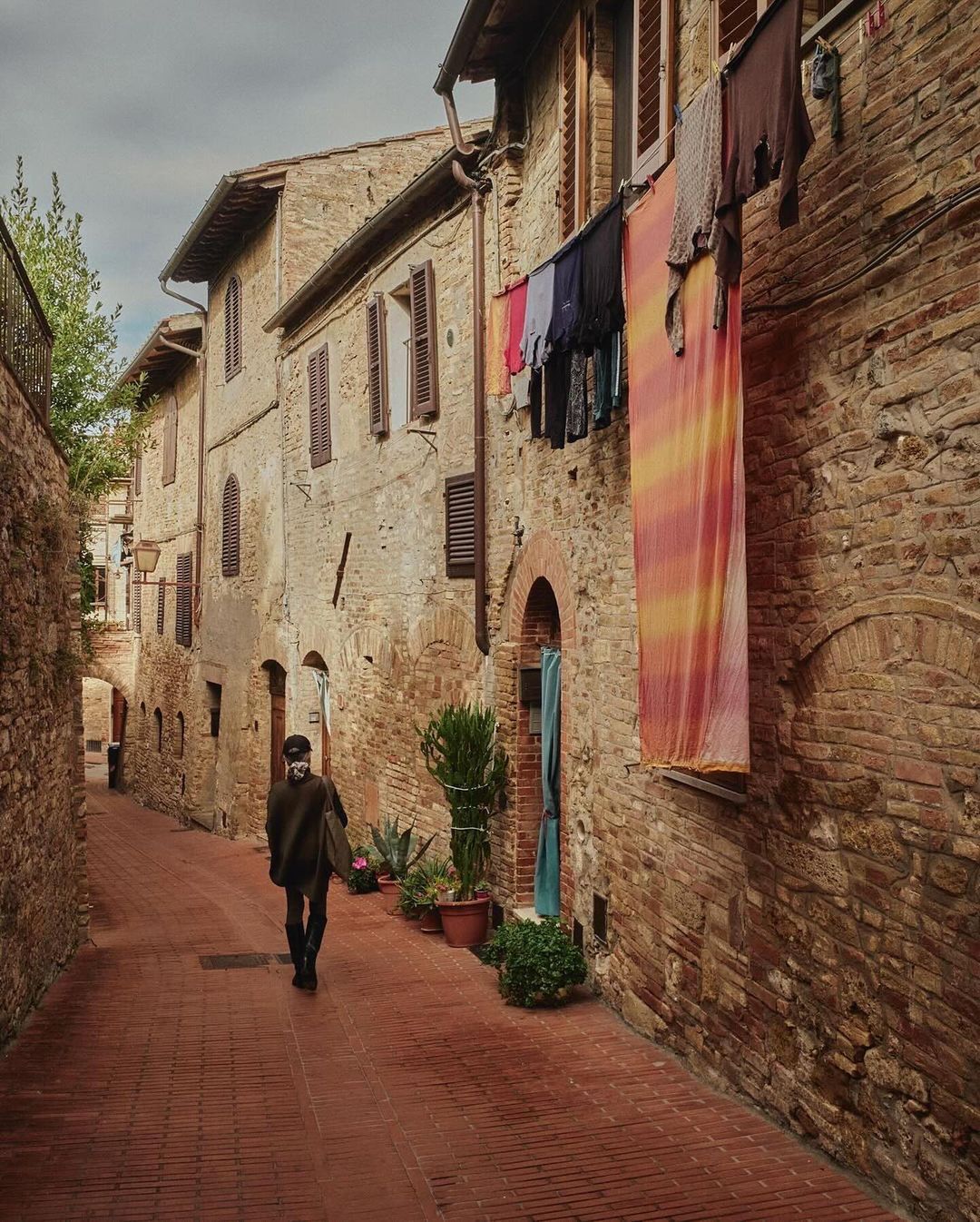 A person walks along a brick road in a rustic Italian town, passing buildings with clothes hanging outside windows on a cloudy day.