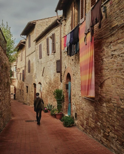 A person walks along a brick road in a rustic Italian town, passing buildings with clothes hanging outside windows on a cloudy day.