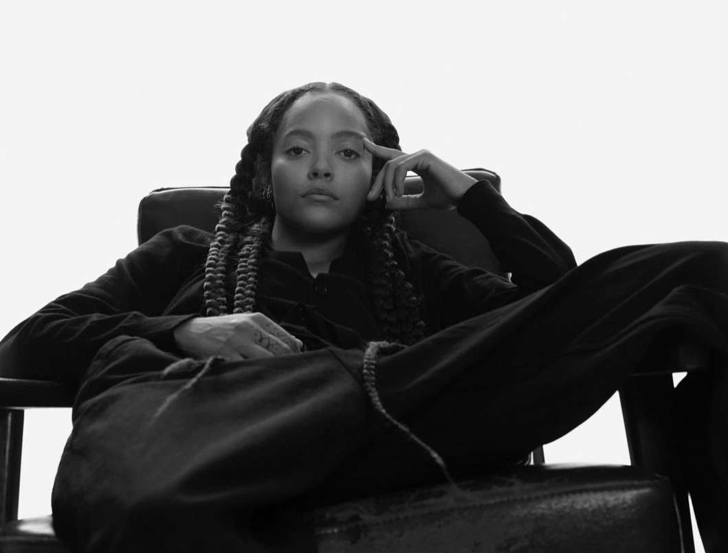 Black and white portrait of a woman with braided hair sitting in a leather armchair against a bright white backdrop, looking at camera.