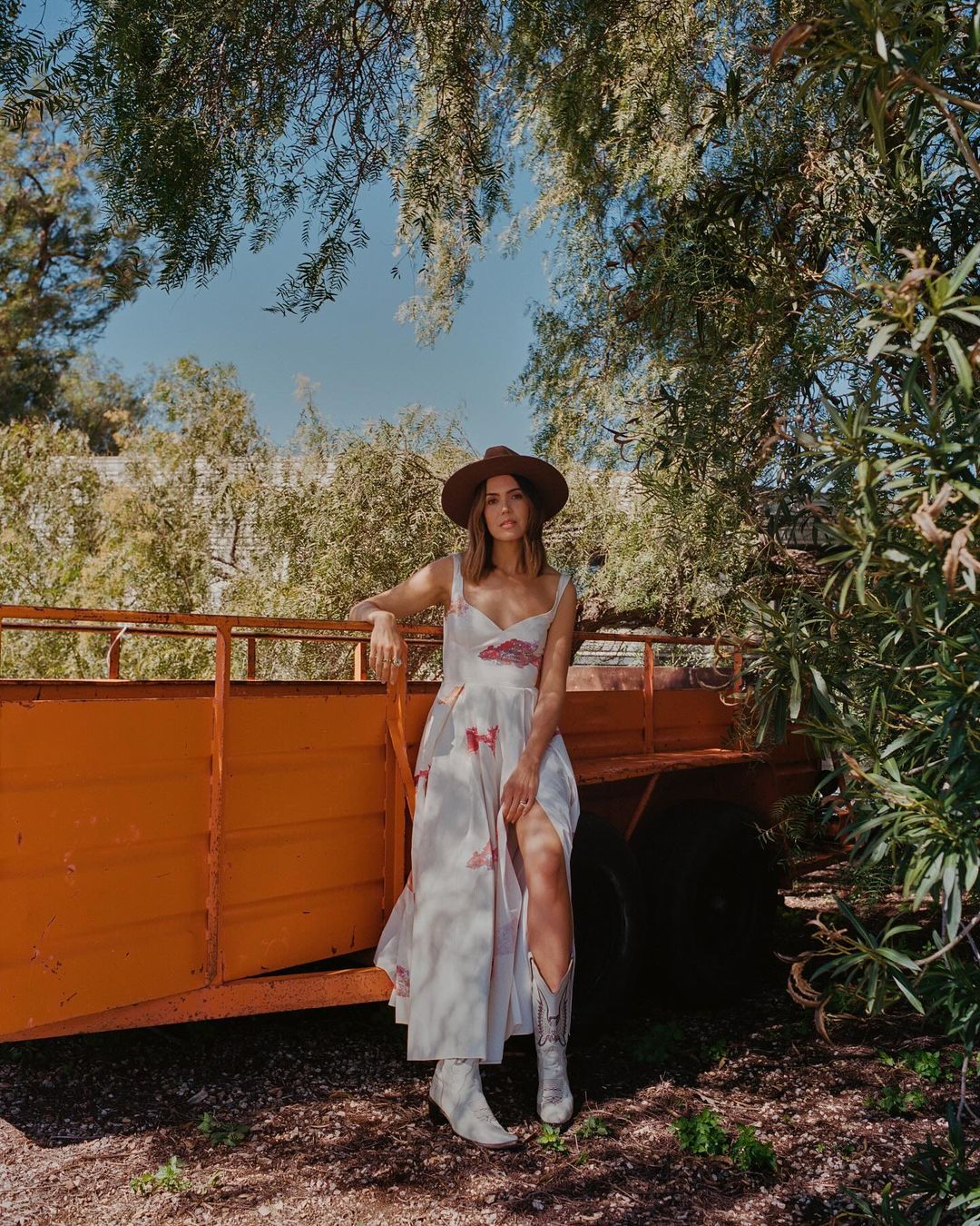 Woman in white dress and cowboy hat leaning against an orange trailer outdoors