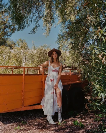 Woman in white dress and cowboy hat leaning against an orange trailer outdoors