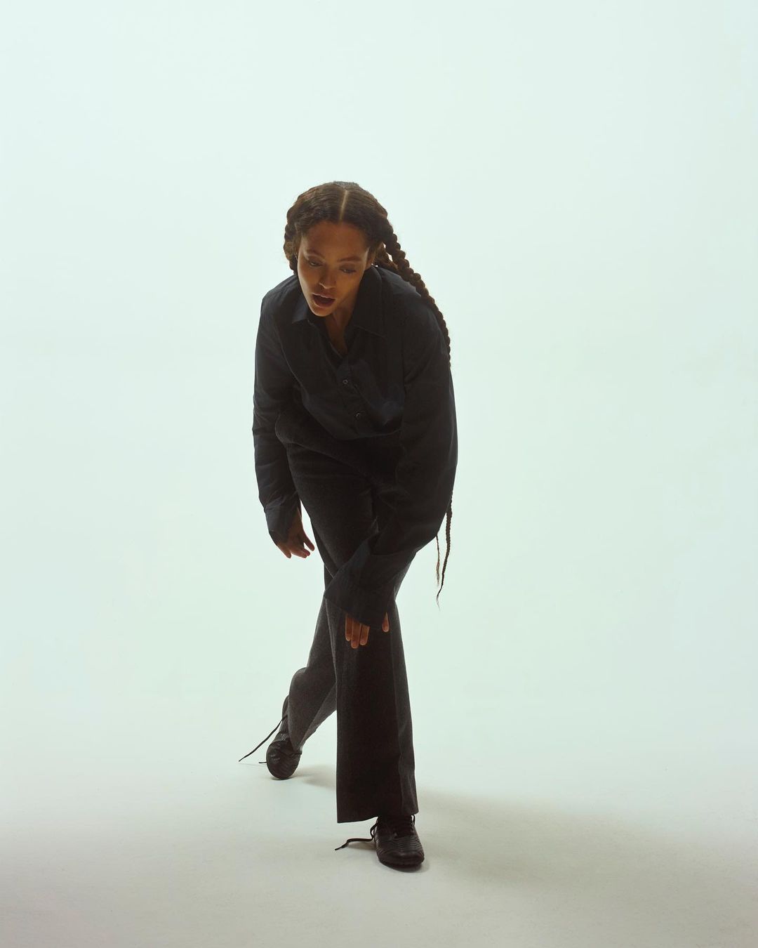A woman in a dark top and trousers poses in a white studio, bending at the waist with her hand on her knee, showcasing a dynamic stance.