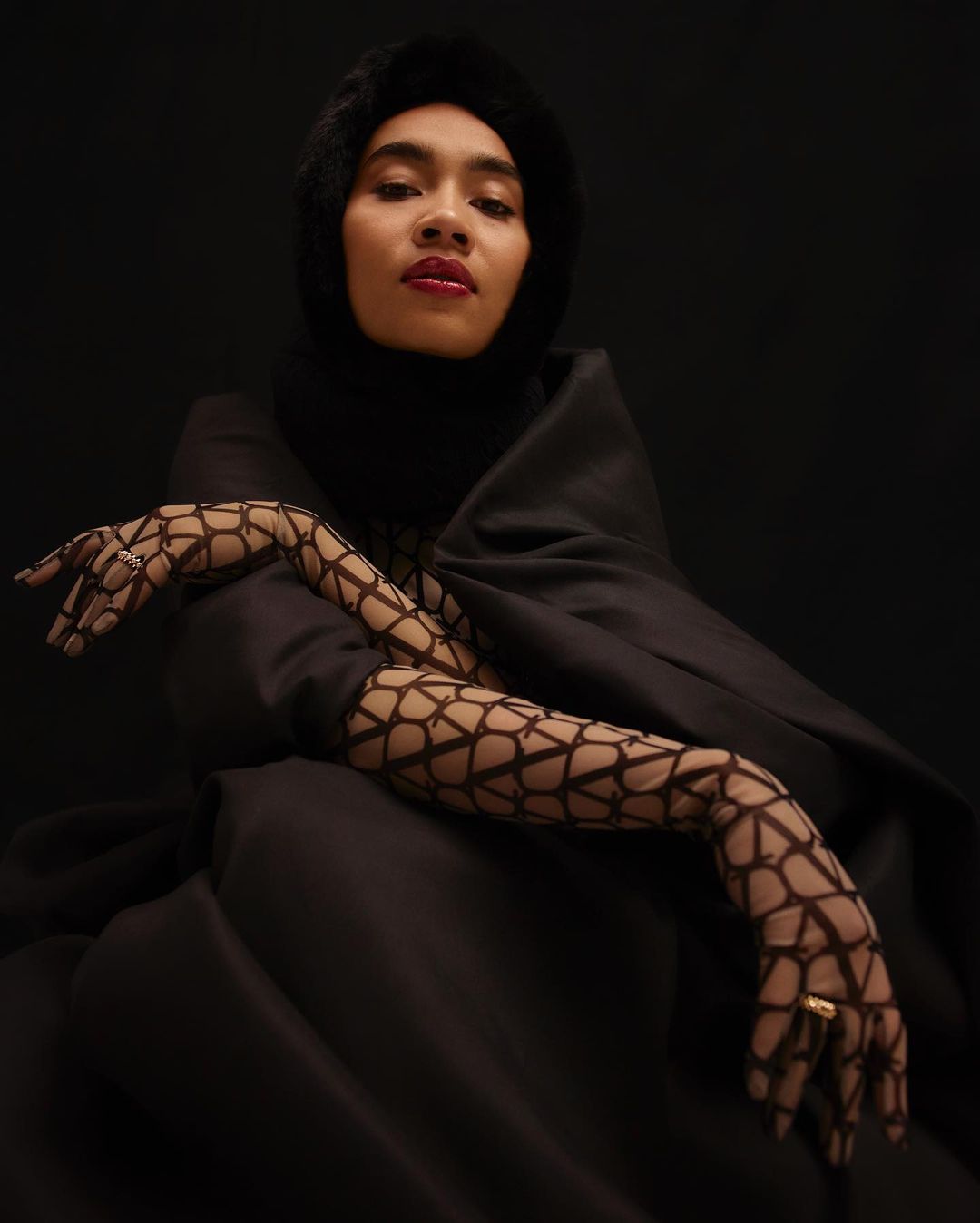 Portrait of a person in a black fur-lined hood and draped garment, wearing patterned net gloves, set against a dark background.