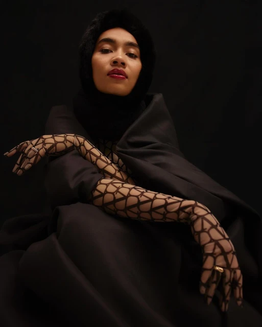 Portrait of a person in a black fur-lined hood and draped garment, wearing patterned net gloves, set against a dark background.