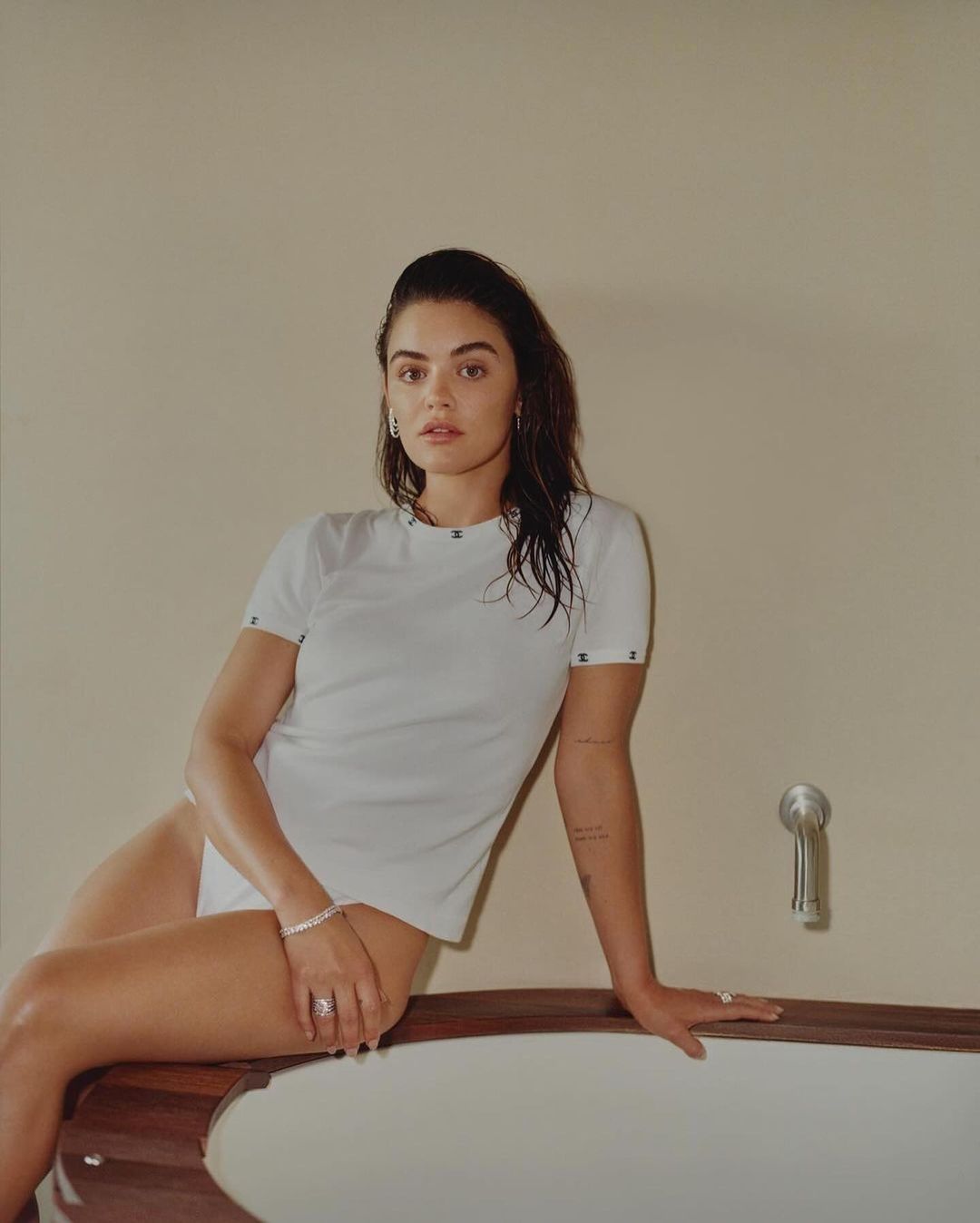 A woman with wet hair, wearing a white t-shirt and underwear, sits on the edge of a bathtub in an indoor setting.
