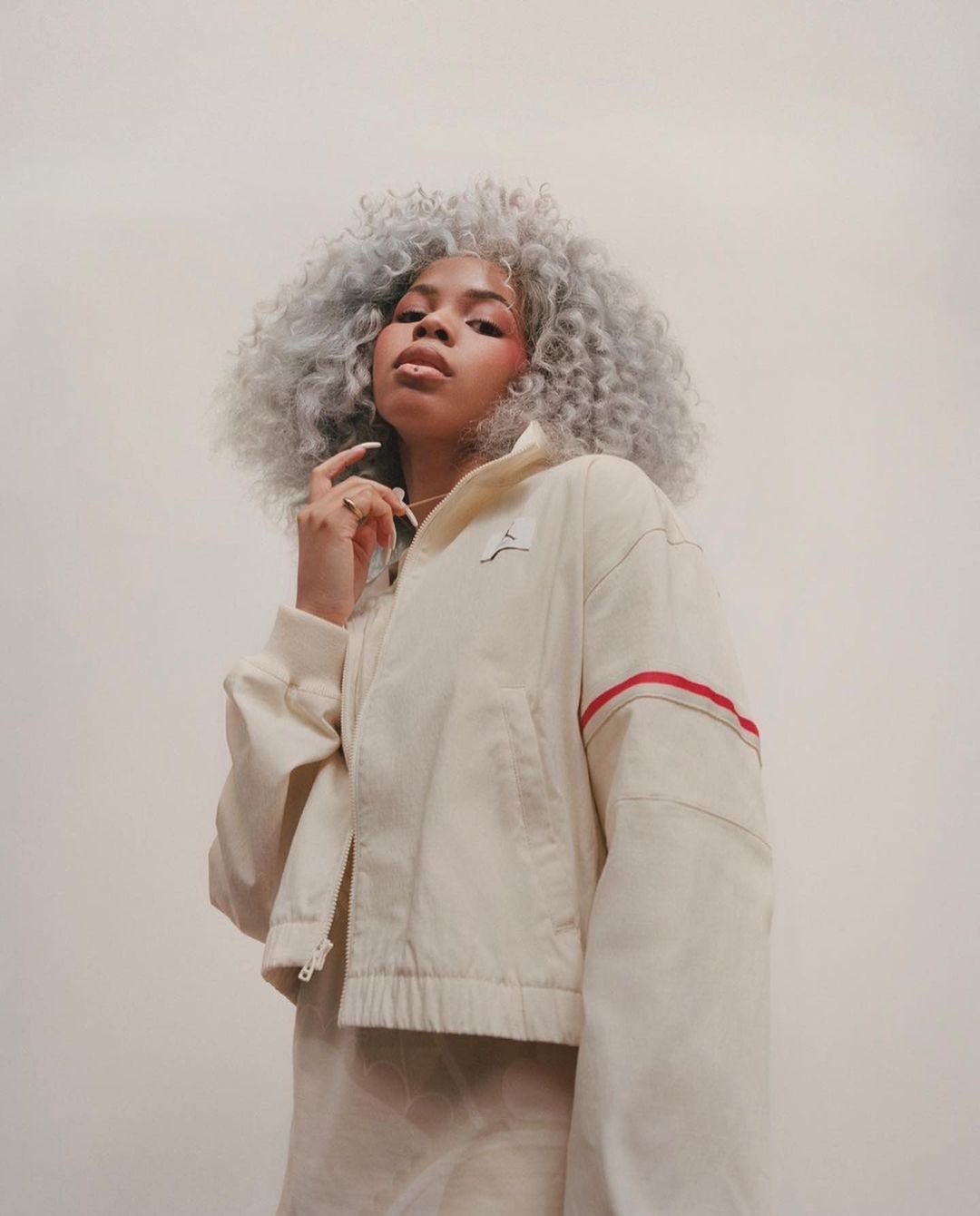 A portrait captures a fashionable person with grey hair, wearing a cream jacket, against a neutral backdrop in a vertical composition.