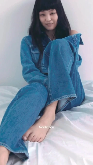 A young adult female sits in a casual pose on a white sheet, wearing a blue denim jacket and wide-leg jeans in a softly lit indoor shot.