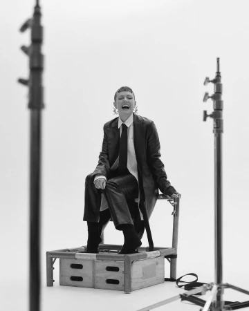 Model with short hair laughing while seated on a crate in a studio, framed by light stands against a white background
