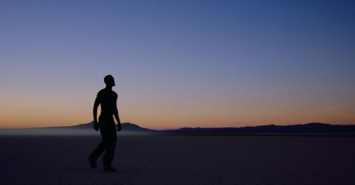 Silhouette of an athletic man walking in a desert landscape during sunset with mountains visible in the distance and clear sky.