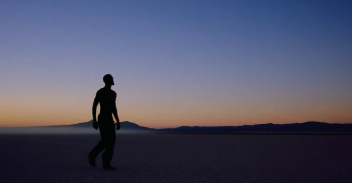 Silhouette of an athletic man walking in a desert landscape during sunset with mountains visible in the distance and clear sky.