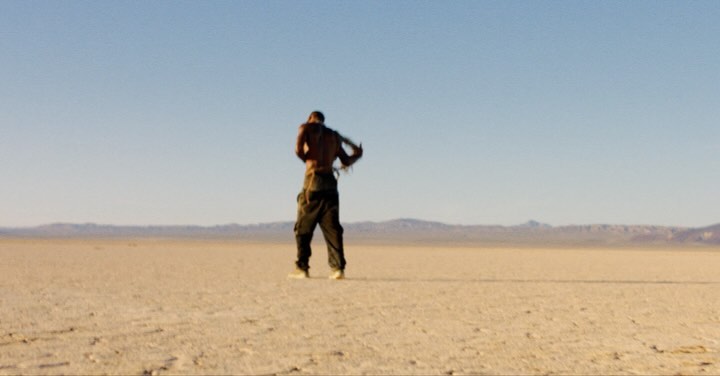 Bare-chested adult in cargo pants stands in arid desert landscape beneath a blue sky with distant mountains on the horizon.