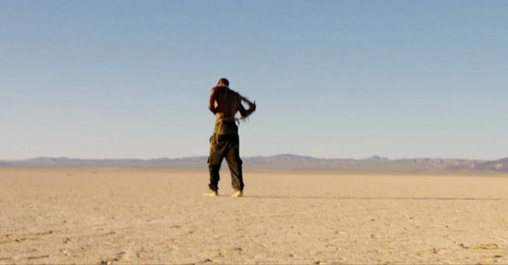 Bare-chested adult in cargo pants stands in arid desert landscape beneath a blue sky with distant mountains on the horizon.
