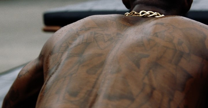 Close-up on the back of a muscular, tattooed Black man wearing a gold chain necklace, against a blurred grey background.
