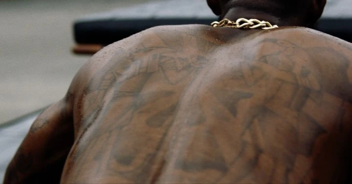 Close-up on the back of a muscular, tattooed Black man wearing a gold chain necklace, against a blurred grey background.