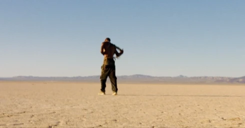 Man standing shirtless with dark trousers in a wide open desert landscape under a clear sky.