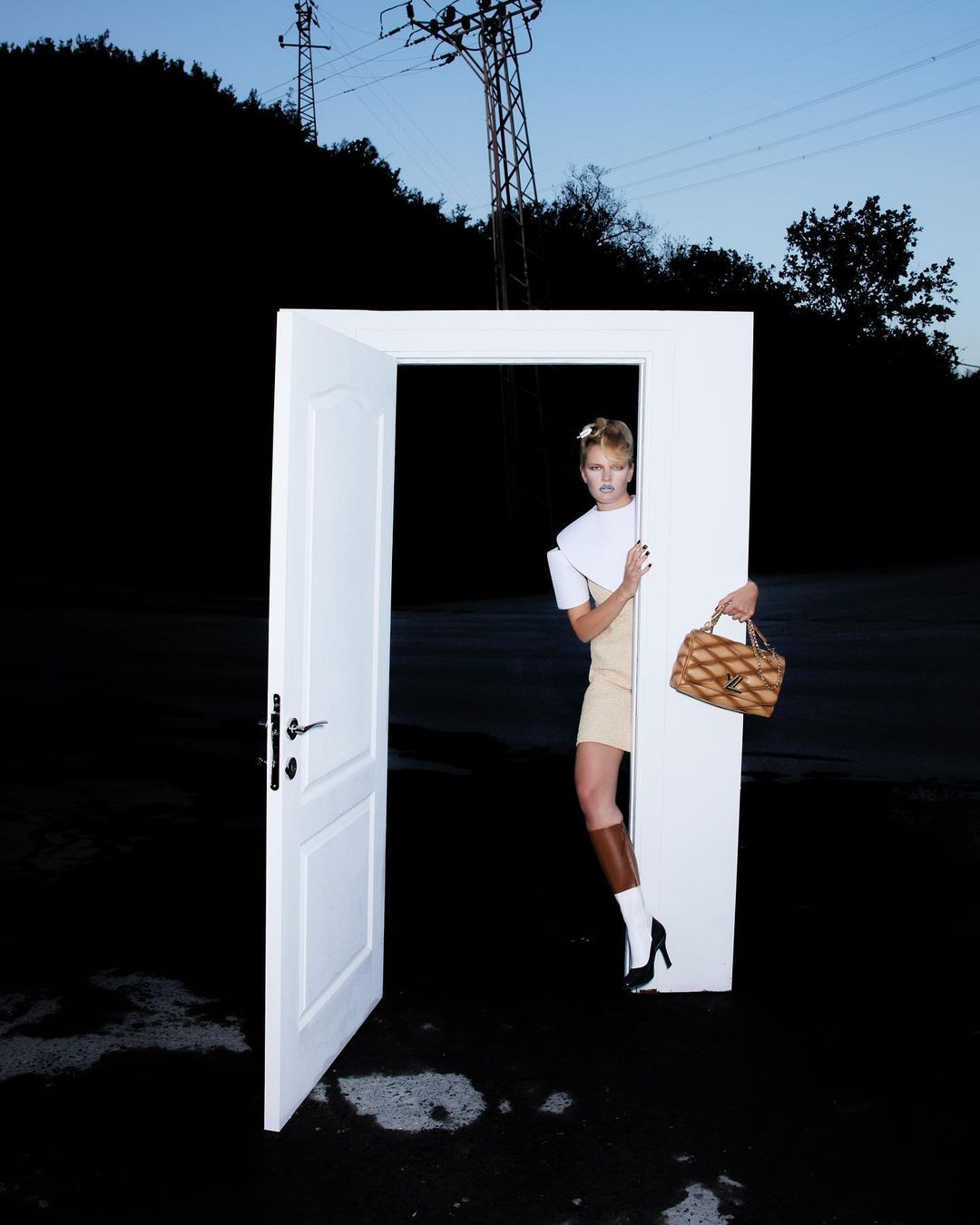 Fashionable woman standing in a doorway in outdoor setting with handbag, wearing modern top, skirt, boots, and high heels.