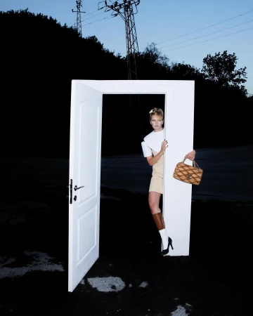 Fashionable woman standing in a doorway in outdoor setting with handbag, wearing modern top, skirt, boots, and high heels.