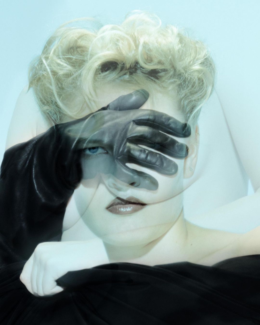 Artistic portrait with a woman's face partially obscured by a gloved hand, set against a light blue background.