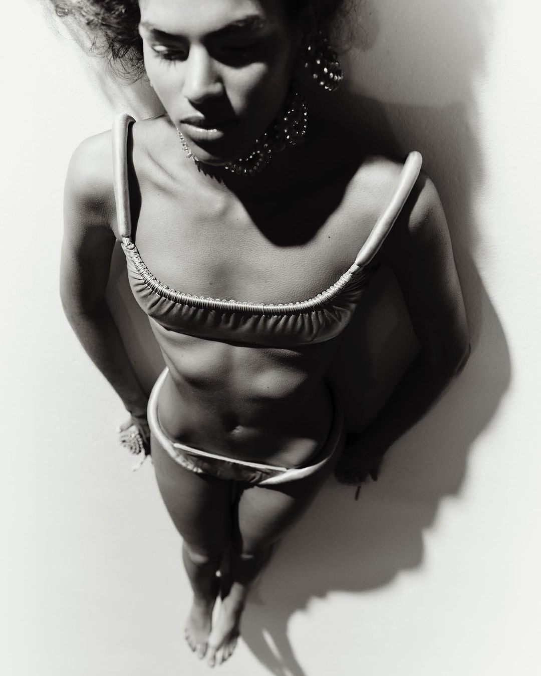Monochrome portrait of a young woman in a bikini, posing with jewelry against a light wall, shot from an overhead angle in soft lighting.