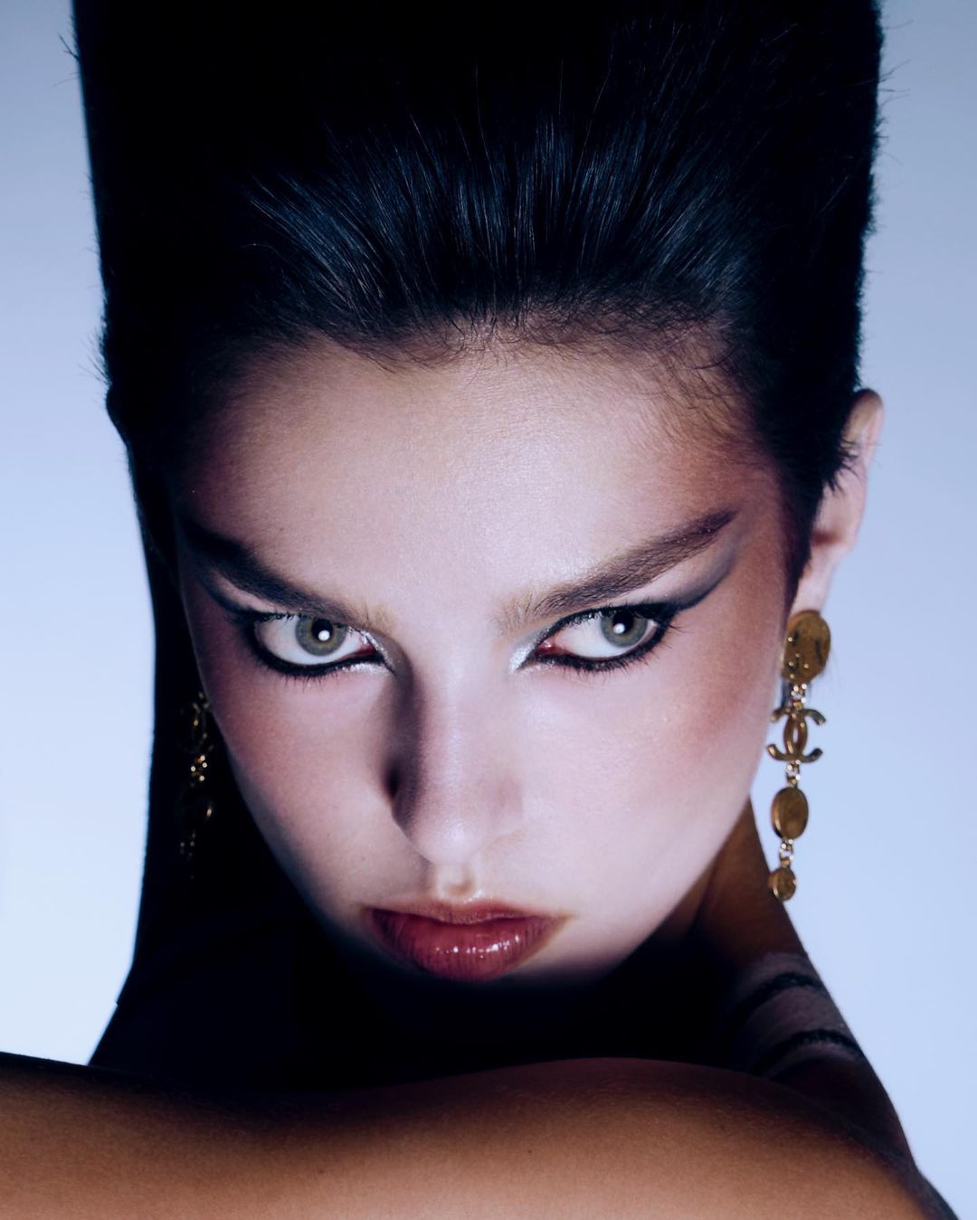Close-up portrait of a young adult woman with dramatic eyeliner and dark, stylized hair, wearing gold Chanel drop earrings in studio lighting.