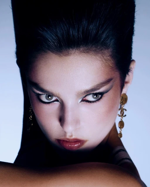 Close-up portrait of a young adult woman with dramatic eyeliner and dark, stylized hair, wearing gold Chanel drop earrings in studio lighting.