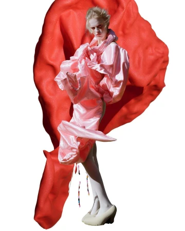 Fashionable woman in pink dress and white tights poses with red fabric in a studio setting, showcasing avant-garde style and design.
