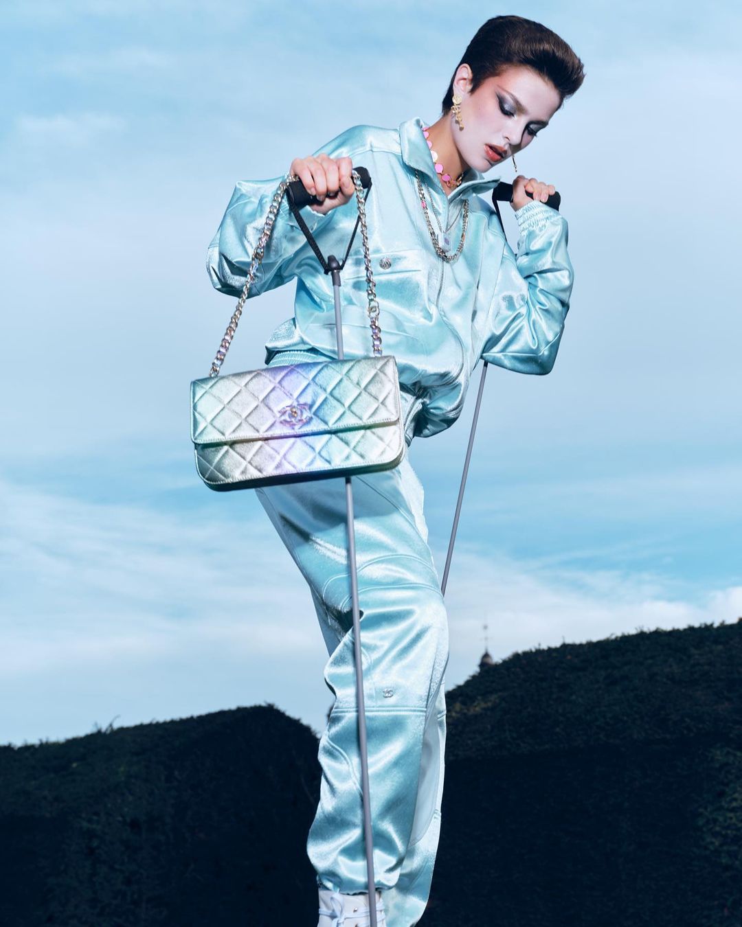 Woman in a light blue tracksuit poses with a Chanel handbag against a sky and dark hill backdrop, creating a fashion-forward image.