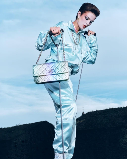 Woman in a light blue tracksuit poses with a Chanel handbag against a sky and dark hill backdrop, creating a fashion-forward image.